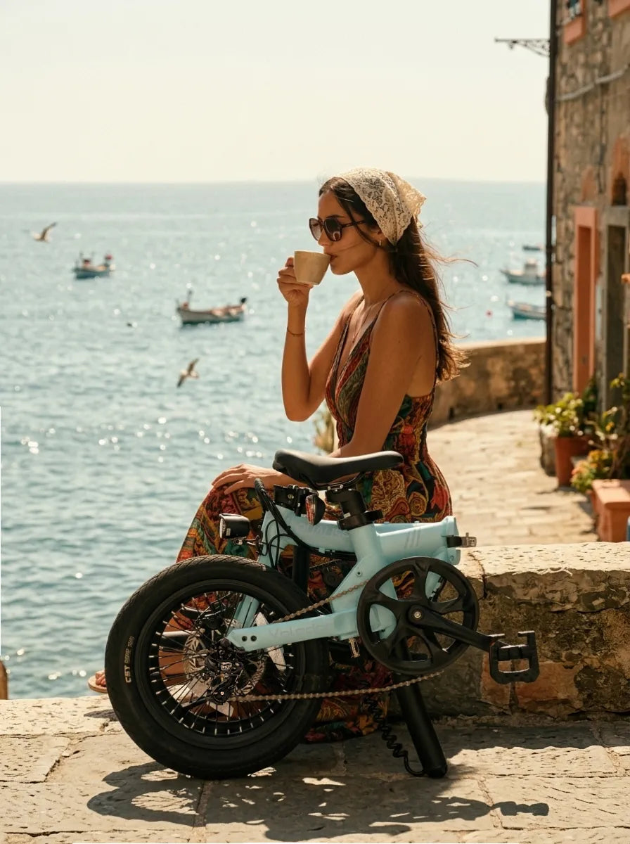 Woman sitting on a stone wall by the sea, drinking coffee with a compact folded bicycle beside her.