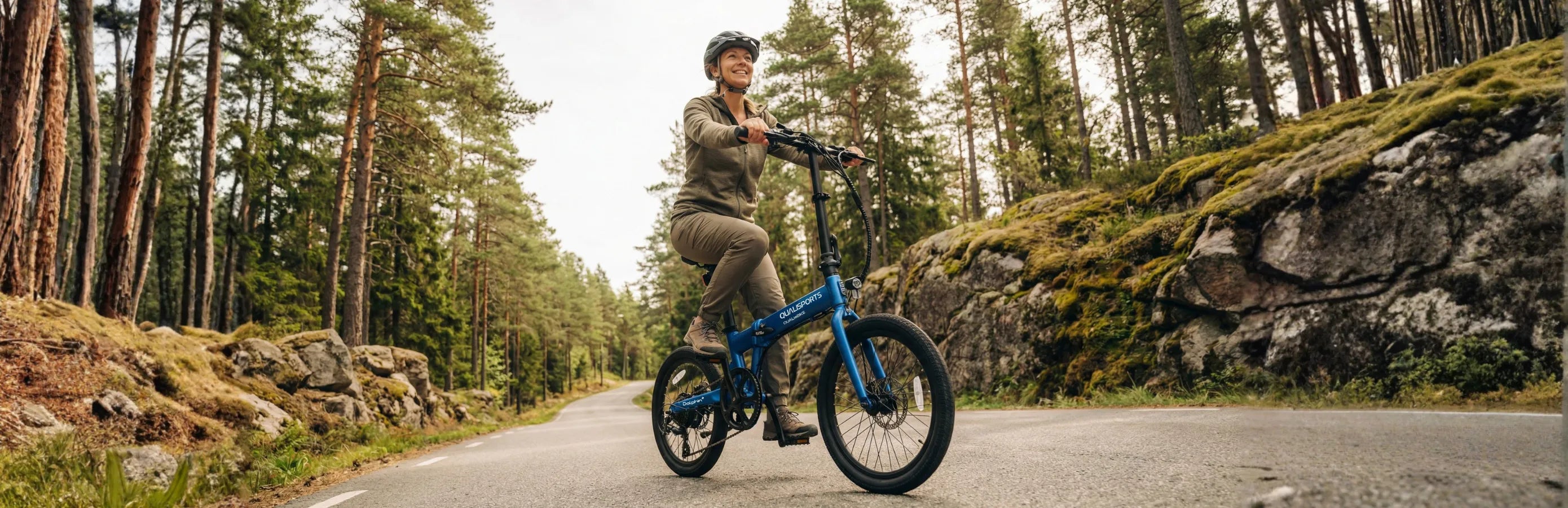 Person riding a blue travel electric bicycle on a forest road with trees and rocks in the background